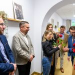 Grupa ludzi w eleganckich strojach rozdaje czerwone róże w jasnym pomieszczeniu. Na ścianie wiszą obrazy i półka z rośliną. Tło jest jasne z żółtymi balonami.