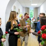 Grupa osób stoi w korytarzu z białymi ścianami, trzymając czerwone róże. Kobiety w czerni, mężczyzna w brązowej marynarce. W tle widać okno i balon.