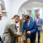 Grupa ludzi w eleganckich strojach w jasnym korytarzu, mężczyzna w granatowym garniturze wita się z osobą w szarej marynarce. Obrazy wiszą na białych ścianach. Balon i prezenty dodają uroczystego charakteru