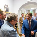 Grupa ludzi w eleganckiej odzieży w jasnym pomieszczeniu; jeden mężczyzna w niebieskim garniturze ściska rękę innemu. W tle białe ściany i obrazy. Kobieta trzyma czerwone róże.