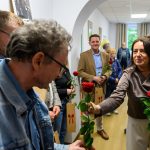 Kobieta wręcza czerwoną różę mężczyźnie w dżinsowej kurtce. Wąski korytarz, jasne ściany, kilka osób trzyma kwiaty. Na ścianach ozdoby i rośliny.
