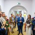 Grupa ludzi w formalnych ubraniach stoi w jasnym korytarzu. Większość trzyma czerwone róże i torebki. Ściany są białe, a podłoga jasnobrązowa.