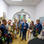 Grupa ludzi stoi w korytarzu, trzymając róże i torby. Ściany są jasne, a na stole widać lawendę. W tle znajdują się balony i wiszące zdjęcia.