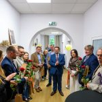 Grupa ludzi w korytarzu z drewnianą podłogą trzyma czerwone róże. Ściany są białe, a w tle widoczne są rośliny i balony. Osoby są elegancko ubrane, stoją w półkolu, patrząc w stronę kamery