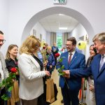 Grupa osób w eleganckich strojach w jasnym wnętrzu wymienia róże i uśmiechy. Tło z jasnymi ścianami i obrazami. Obecne są balony dekoracyjne.