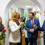 Grupa ludzi w eleganckich strojach stoi w jasnym pomieszczeniu, trzymając czerwone róże. Tło z białymi ścianami i obrazkami. Uśmiechy sugerują uroczyste wydarzenie.