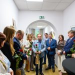 Na zdjęciu widoczna jest grupa osób stojących w korytarzu i trzymających czerwone róże. W tle jasne ściany i okno, tło jest stonowane. Na środku osoby, jedna w dżinsowej kurtce.