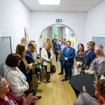 Na zdjęciu grupa osób w korytarzu, ubranych elegancko, trzyma czerwone róże. Jasne ściany i drewniana podłoga. Nastroje są radosne.