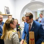 Na zdjęciu widać grupę ludzi podczas spotkania w jasnym pomieszczeniu. Osoba w niebieskiej marynarce wymienia uścisk dłoni z kobietą o blond włosach, oboje trzymają torby. W tle inni uczestnicy trzymają czerwone róże i patrzą z u