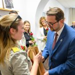 Grupa ludzi w eleganckich strojach wymienia upominki w jasnym pokoju. Kobieta trzyma różę i prezent. Mężczyzna w niebieskiej marynarce wręcza pakiet.