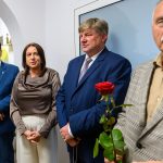 Grupa ludzi stoi w jasnym pomieszczeniu, większość w eleganckich ubraniach. Jedna osoba trzyma czerwoną różę. Tło ma biały kolor z niebieskim elementem.