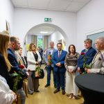 Grupa osób stoi w korytarzu, trzymając czerwone róże. Tło jest jasne, z białymi ścianami i jasną podłogą. Na stoliku obok jest tabliczka z napisem “Mazowsze”.
