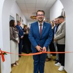 Mężczyzna w niebieskim garniturze przecina czerwoną wstęgę. Stoi w jasnym korytarzu, wokół inni formalnie ubrani ludzie. W tle białe ściany i kwiatowa dekoracja.