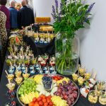 Stół z kolorowymi przekąskami owocowymi i deserami w słoiczkach na czarnym obrusie. W tle zielone kwiaty w wazonie. Ludzie stoją obok.