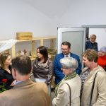 Grupa ludzi rozmawia w jasnym pomieszczeniu z półkami. Ubrania są kolorowe, dominuje beż i szary. W tle dekoracje i drewniane kosze.