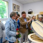 Grupa osób w jasnym pomieszczeniu ogląda otwarty piec ceramiczny. Tło stanowi ściana z oknem i obrazami. Niektórzy trzymają czerwone róże.