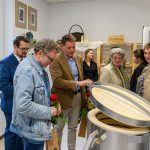 Grupa osób stoi wokół nowoczesnego pieca ceramicznego. Ubrania w stonowanych kolorach, wnętrze jasne. Przez okno widać zielone drzewa.
