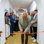 Mężczyzna w beżowej marynarce przecina czerwoną wstęgę. W tle grupa osób w formalnych strojach. Jasne, nowoczesne wnętrze.