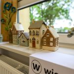 Na białym parapecie stoją małe, ceramiczne domki w pastelowych kolorach. W tle widać drzewa za oknem. Obok znajduje się drewniana dekoracja z kwiatami.