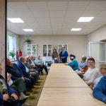 Długi stół w sali z jasnym oświetleniem. Grupa osób siedzi i stoi wokół, z roślinami i regałami w tle. Dominują beże i błękity.