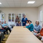 Grupa osób siedzi przy drewnianym stole w jasnym pokoju. W tle regały z książkami i ozdobami. Ściany w beżowym kolorze.