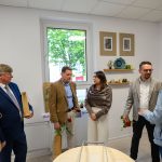 Na zdjęciu widać grupę osób w pomieszczeniu o białych ścianach. Niektórzy z nich trzymają czerwone róże i torby. W tle znajdują się półki z ozdobami.