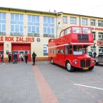 Czerwony piętrowy autobus stoi przed budynkiem straży pożarnej z napisem OSP Dobre Rok Zał. 1915. Grupa ludzi obok pojazdu. Pogodne niebo i brukowane podłoże.