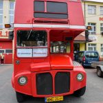 Czerwony piętrowy autobus stoi na brukowanym placu. W tle jasne budynki z dużymi oknami. Rejestracja polska, napis Mazowsze.