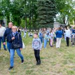 Grupa ludzi na zielonej trawie w parku. Drzewa w tle, jasne ubrania uczestników. Rodzinna atmosfera eventu plenerowego.