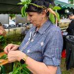 Kobieta w niebieskiej koszuli plecie wianek z zielonych liści. W tle namiot z żółto-zielonym dachem. Na zewnątrz, przy stole.