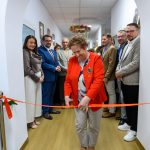 Starsza kobieta w czerwonej marynarce przecina czerwoną wstęgę w jasnym korytarzu, otoczona przez grupę ludzi.