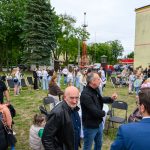Grupa ludzi na trawniku przed budynkiem. Zielone drzewo z lewej, namiot z prawej. Dzieci i dorośli rozmawiają.