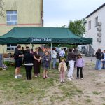 Na zdjęciu widoczny jest zielony namiot z napisem Gmina Dobre, otoczony ludźmi. Tło stanowi jasny budynek z oknami i trawnikiem. Grupa osób rozmawia i spaceruje na zewnątrz.