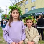 Dwie dziewczynki w wiankach z liści stoją na tle zielonego namiotu i żółtego budynku. Jedna ma fioletowy sweter, druga beżową kurtkę. W tle grupka osób.