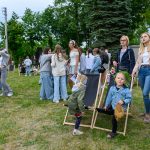 Grupa ludzi na zielonej trawie; dzieci, młodzież i dorośli w casualowych ubraniach, w tle drzewa i niebo.