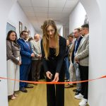 Kobieta w czarnym stroju przecina czerwoną wstęgę w białym korytarzu. W tle grupa elegancko ubranych osób. Wszystko odbywa się w jasnym, przestronnym pomieszczeniu.