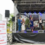 Na scenie występują cztery osoby, w tle zestaw perkusyjny. Na pierwszym planie biały baner z czerwonym logo Mazowsze. Tło stanowi nowoczesny budynek z zielenią.