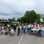 Na zdjęciu pokazane jest wydarzenie plenerowe z dużą grupą ludzi na placu. W tle są drzewa, namioty i budynek. Przeważają kolory szary, zielony i biały.