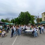 Na zdjęciu widać tłum ludzi podczas wydarzenia plenerowego. Przedstawione są kolorowe namioty, dzieci na leżakach i zielone drzewa w tle. Po prawej stronie znajduje się budynek o żółtych ścianach.