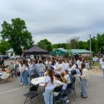 Na zdjęciu widać grupę ludzi biorących udział w plenerowym wydarzeniu. Tło stanowią zielone drzewa i namioty. Uczestnicy są w swobodnych ubraniach, atmosfera jest radosna.