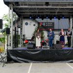 Scena plenerowa z ludźmi przemawiającymi do mikrofonów. Tło z banerem Powiat Miński. Po lewej biały roll-up Mazowsze.