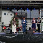 Grupa osób stoi na scenie podczas wydarzenia powiatowego. W tle widoczny jest baner z napisem Powiat Miński. Na scenie znajdują się instrumenty muzyczne i sprzęt audio.