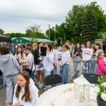 Grupa młodych ludzi na zewnątrz z zielonymi i czarnymi namiotami w tle. Ubrani na sportowo, pogoda jest pochmurna. Na przodzie biała latarnia na ozdobnym stole.