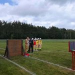 Grupa strażaków w jasnych uniformach stoi na zielonej trawie. W tle jest las i pochmurne niebo. Po lewej widać drewnianą przeszkodę i sprzęt strażacki.