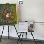 Obraz przedstawia bukiet kolorowych kwiatów w wazonie na tle ciemnej zieleni. Obok umieszczono tabliczkę z informacjami na białym tle. Całość ustawiona jest na sztalugach w jasnym pomieszczeniu.
