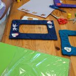 Kolorowe warsztaty plastyczne na drewnianym stole. Dzieci ozdabiają ramki wycinankami i klejem. Na stole zielony papier, nożyczki, klej.