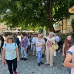 Grupa osób spaceruje po brukowanej ulicy, częściowo zacieniona przez drzewa. W tle widać budynki i zadaszenia. Ubrania w jasnych i ciemnych kolorach.