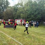Strażacy w hełmach biegną po zielonym polu. W tle widoczne wozy strażackie i drzewa. Scena odbywa się na zewnątrz.