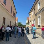 Grupa ludzi stoi na wąskiej, brukowanej uliczce między pastelowymi budynkami. Tło to czyste, niebieskie niebo. Po prawej stronie widoczne są białe okna i czerwony szyld.