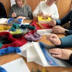 Dwie osoby siedzą przy stole i pracują z kolorową wełną na filcowaniu. Wokół leżą różnobarwne pasma: czerwone, niebieskie, żółte. W tle widać drewnianą szafkę i zieloną roślinę.